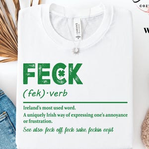 May include: White t-shirt with the word "FECK" in green, featuring a shamrock. The definition of "FECK" is printed below, describing it as an Irish expression of annoyance. The shirt also includes the text "Elegant Cozy Tees" and "WHITE".