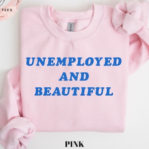 May include: A light pink sweatshirt with the words "UNEMPLOYED AND BEAUTIFUL" in blue block letters. The word "PINK" is printed in black at the bottom of the image. The sweatshirt is from Elegant Cozy Tees.