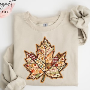 May include: Sand-colored sweatshirt featuring a large, detailed leaf design. The leaf is composed of various plaid and autumn-themed patterns in shades of red, green, and orange. The word "SAND" is printed at the bottom. The brand name "Elegant Cozy Tees" is in the upper left corner.