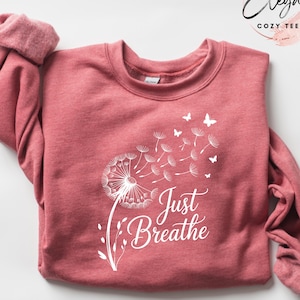 Peut inclure: Un sweat-shirt &agrave; manches longues rouge &eacute;carlate chin&eacute; avec un motif blanc repr&eacute;sentant un pissenlit dont les graines s'envolent et de petits papillons. Le texte Just Breathe est imprim&eacute; en &eacute;criture blanche sous le motif du pissenlit.