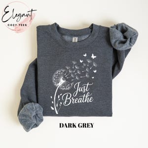 Peut inclure: Un sweat-shirt &agrave; manches longues gris fonc&eacute; avec une impression graphique blanche repr&eacute;sentant un pissenlit dont les graines s'envolent au vent, de petits papillons et le texte Just Breathe en &eacute;criture cursive. Col rond et poignets c&ocirc;tel&eacute;s.