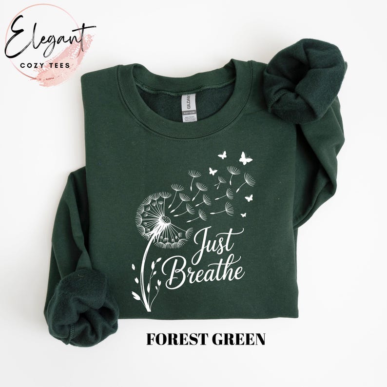 Peut inclure: Un sweat-shirt &agrave; manches longues vert for&ecirc;t avec un motif blanc repr&eacute;sentant un pissenlit dont les graines se transforment en papillons. Le texte "Just Breathe" est imprim&eacute; en police cursive blanche sous le motif sur le devant.
