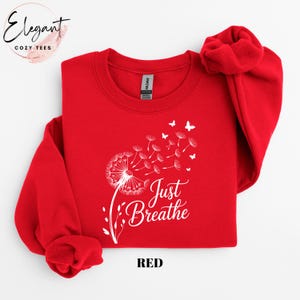 Peut inclure: Un sweat-shirt rouge vif &agrave; manches longues et col rond, orn&eacute; d'un motif blanc repr&eacute;sentant un pissenlit dont les graines se transforment en petits papillons. Le texte Just Breathe est imprim&eacute; en lettres cursives blanches.
