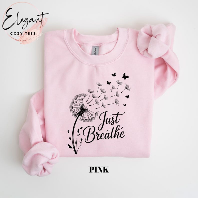 Peut inclure: Un sweat-shirt &agrave; col rond rose clair avec un motif noir repr&eacute;sentant un pissenlit dont les graines s'envolent pour devenir de petits papillons. Le texte Just Breathe est imprim&eacute; en police cursive noire sous le motif sur le devant.