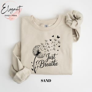 Peut inclure: Un sweat-shirt &agrave; col rond couleur sable avec un motif noir repr&eacute;sentant un pissenlit dont les graines s'envolent en se transformant en petits papillons. Le texte "Just Breathe" est imprim&eacute; en noir sous le motif sur le devant.