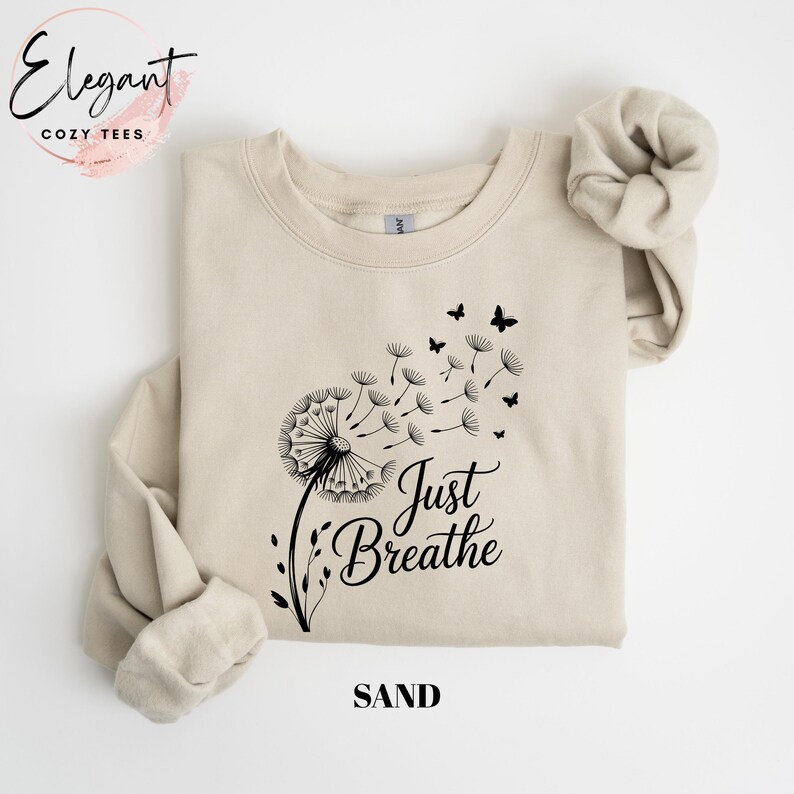 Peut inclure: Un sweat-shirt &agrave; col rond couleur sable avec un motif noir repr&eacute;sentant un pissenlit dont les graines s'envolent en se transformant en petits papillons. Le texte "Just Breathe" est imprim&eacute; en noir sous le motif sur le devant.