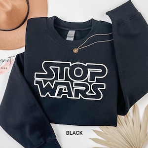 May include: A black long-sleeve crewneck sweatshirt featuring the white text STOP WARS printed in a stylized font across the chest. The garment has a ribbed neckline, cuffs, and hem, with a Gildan brand label visible inside the collar.