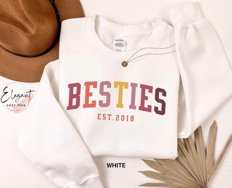May include: A white long-sleeve crewneck sweatshirt featuring the word BESTIES in a multi-colored collegiate font with the text EST. 2018 printed underneath in a smaller coral font. The garment has a ribbed collar, cuffs, and hemline.