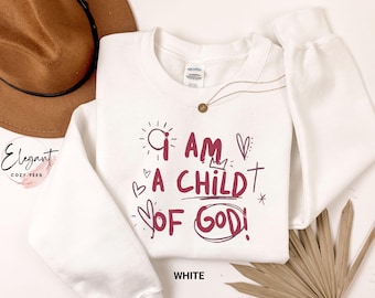 May include: A white long-sleeve crewneck sweatshirt featuring the maroon text "I AM A CHILD OF GOD!" printed on the front. The design includes hand-drawn style graphics of a sun, a small crown, a cross, and several hearts surrounding the lettering.