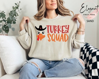 May include: A sand-coloured crewneck sweatshirt featuring a graphic of a turkey wearing a black cowboy hat with a red star. The text "TURKEY SQUAD" is printed in red and orange lettering. The sweatshirt has long sleeves and a ribbed neckline.