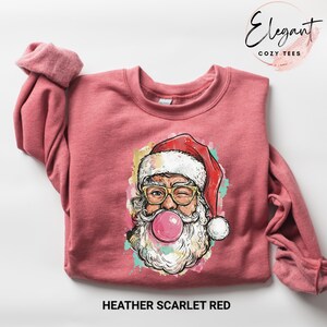 May include: Heather scarlet red sweatshirt featuring a graphic of Santa Claus winking and blowing a bubblegum bubble. Santa is wearing gold-rimmed glasses and a red hat. The sweatshirt has a crew neck and long sleeves. The text "Elegant Cozy Tees" is in the upper right corner.