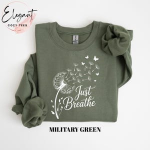 Peut inclure: Un sweat-shirt &agrave; col rond vert militaire avec un motif blanc repr&eacute;sentant un pissenlit dont les graines s'envolent en petits papillons. Le texte Just Breathe est imprim&eacute; en police cursive blanche sous le motif sur le devant.