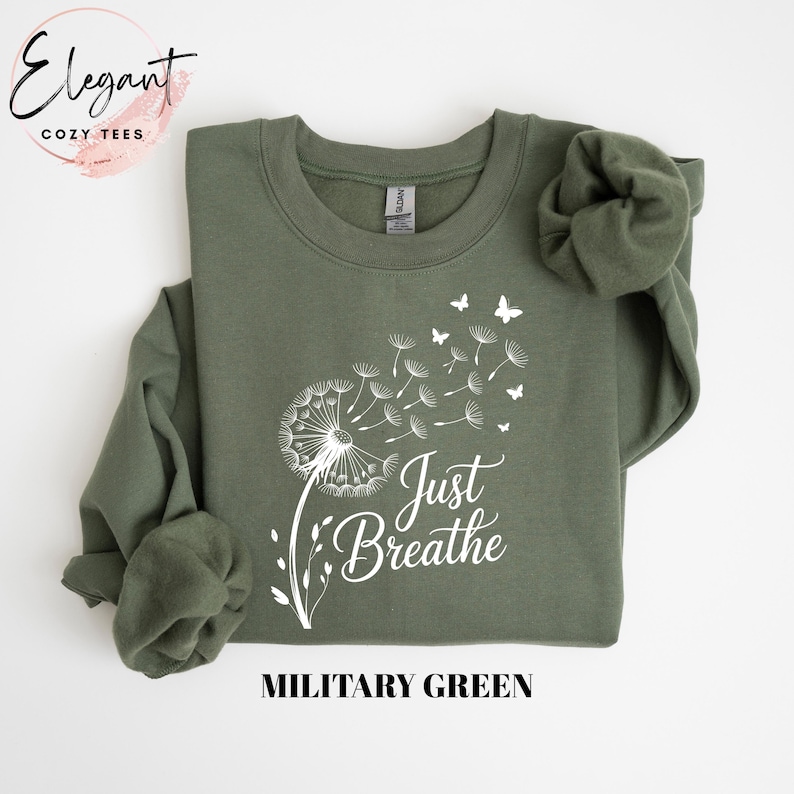 Peut inclure: Un sweat-shirt &agrave; col rond vert militaire avec un motif blanc repr&eacute;sentant un pissenlit dont les graines s'envolent en petits papillons. Le texte Just Breathe est imprim&eacute; en police cursive blanche sous le motif sur le devant.