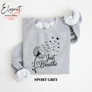 Peut inclure: Un sweat-shirt &agrave; col rond gris sport avec un motif noir repr&eacute;sentant un pissenlit dont les graines s'envolent en se transformant en petits papillons. Le texte Just Breathe est imprim&eacute; en police cursive noire sous le pissenlit.