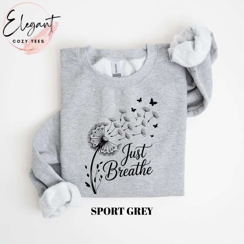 Peut inclure: Un sweat-shirt &agrave; col rond gris sport avec un motif noir repr&eacute;sentant un pissenlit dont les graines s'envolent en se transformant en petits papillons. Le texte Just Breathe est imprim&eacute; en police cursive noire sous le pissenlit.