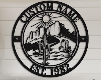 May include: A black metal wall art piece with a desert landscape design, including cacti, mountains, and a sun. The text "CUSTOM NAME" and "EST. 1982" are displayed on the circular frame. The artwork is mounted on a white wall above a brown leather sofa.