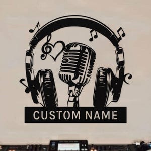 以下が含まれることがあります： DJをテーマにした黒い金属製の壁掛けアート。ヘッドフォン、音符、マイク、ハートのデザイン。テキスト「CUSTOM NAME」がプレートに表示されています。下には、ターンテーブルとミキサーを備えたDJセットアップがあります。