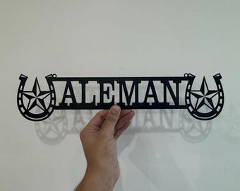 May include: A black metal wall sign featuring the name ALEMAN in capital letters, flanked on both sides by a horseshoe design containing a five-pointed star. The piece is constructed from a single flat sheet of metal with cut-out details.