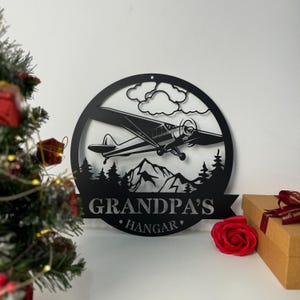 May include: Black metal sign with a silhouette of a small airplane flying over mountains and trees. The sign reads "Grandpa's Hangar".