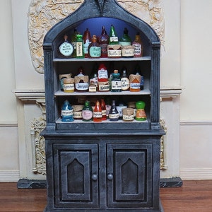 May include: A black wooden cabinet with glass doors and shelves filled with potion bottles. The bottles have labels with names like "Fairy Wine", "Beetle Juice", and "Blood".