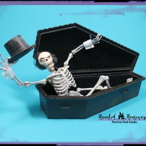 May include: A skeleton wearing a top hat and white cuffs is sitting up in a black coffin. The coffin is open and the skeleton is looking up. The background is a bright blue.