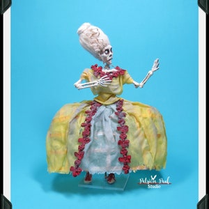 May include: A skeletal figure wearing a yellow dress with red trim and a white wig. The figure is posed with one arm raised and the other holding the dress. The figure is standing on a clear display stand. The image is signed "Patricia Paul Studio".