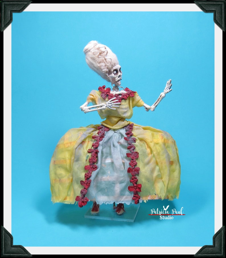 May include: A skeletal figure wearing a yellow dress with red trim and a white wig. The figure is posed with one arm raised and the other holding the dress. The figure is standing on a clear display stand. The image is signed "Patricia Paul Studio".