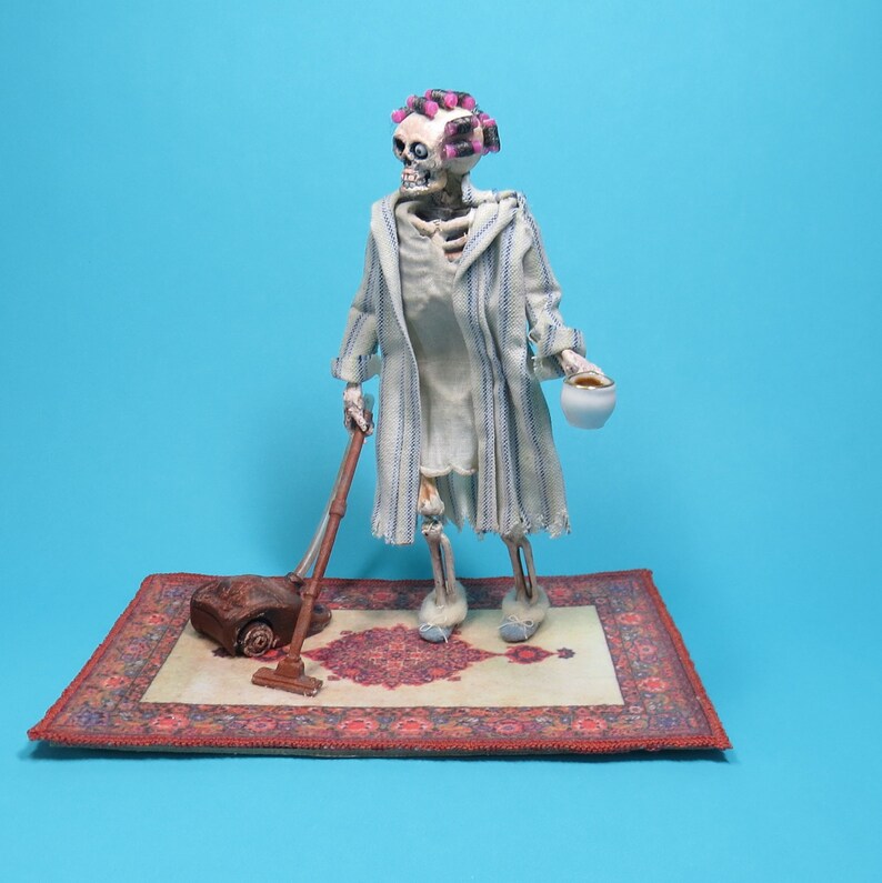 May include: A skeleton doll wearing a white and blue striped robe, pink hair rollers, and slippers, holding a cup and vacuuming a red and cream patterned rug.