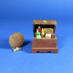 Puede incluir: Un cofre de madera en miniatura con una tapa con bisagras, abierto para revelar una colección de artículos en miniatura, incluyendo botellas, libros y otras baratijas. El cofre está pintado de color marrón oscuro y tiene una manija de latón.