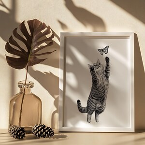 Pode incluir: Uma impress&atilde;o em preto e branco emoldurada mostra um gato malhado alcan&ccedil;ando uma borboleta. O gato est&aacute; sobre as patas traseiras, com as patas dianteiras estendidas. Uma folha marrom em um vaso de vidro e duas pinhas est&atilde;o em primeiro plano.