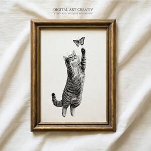 Pode incluir: Uma impress&atilde;o emoldurada em preto e branco com um gato brincalh&atilde;o alcan&ccedil;ando uma borboleta. O gato &eacute; retratado com listras detalhadas, em um fundo neutro. A moldura &eacute; marrom dourado quente. O texto na parte superior diz "DIGITAL ART CREATIV".