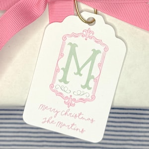 Könnte beinhalten: Ein Geschenkanhänger mit einer rosa Schleife. Der Anhänger ist weiß mit einem rosa und grünen dekorativen Rahmen um den Buchstaben "M". Der Text "Merry Christmas The Martins" ist darunter geschrieben.