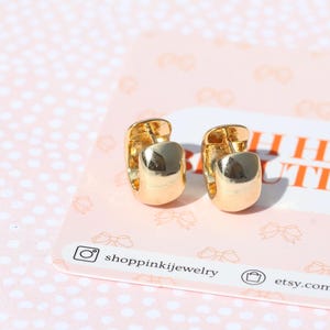 May include: A pair of gold-colored hoop earrings with a rounded, rectangular design. The earrings are displayed on a pink card with the text "OH HEY BEAUTIFUL" and the shop's social media handles.