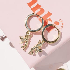 May include: Gold-colored hoop earrings with a charm of a small figure with outstretched arms. The earrings are displayed on a pink surface with the word "Pink" in orange letters.