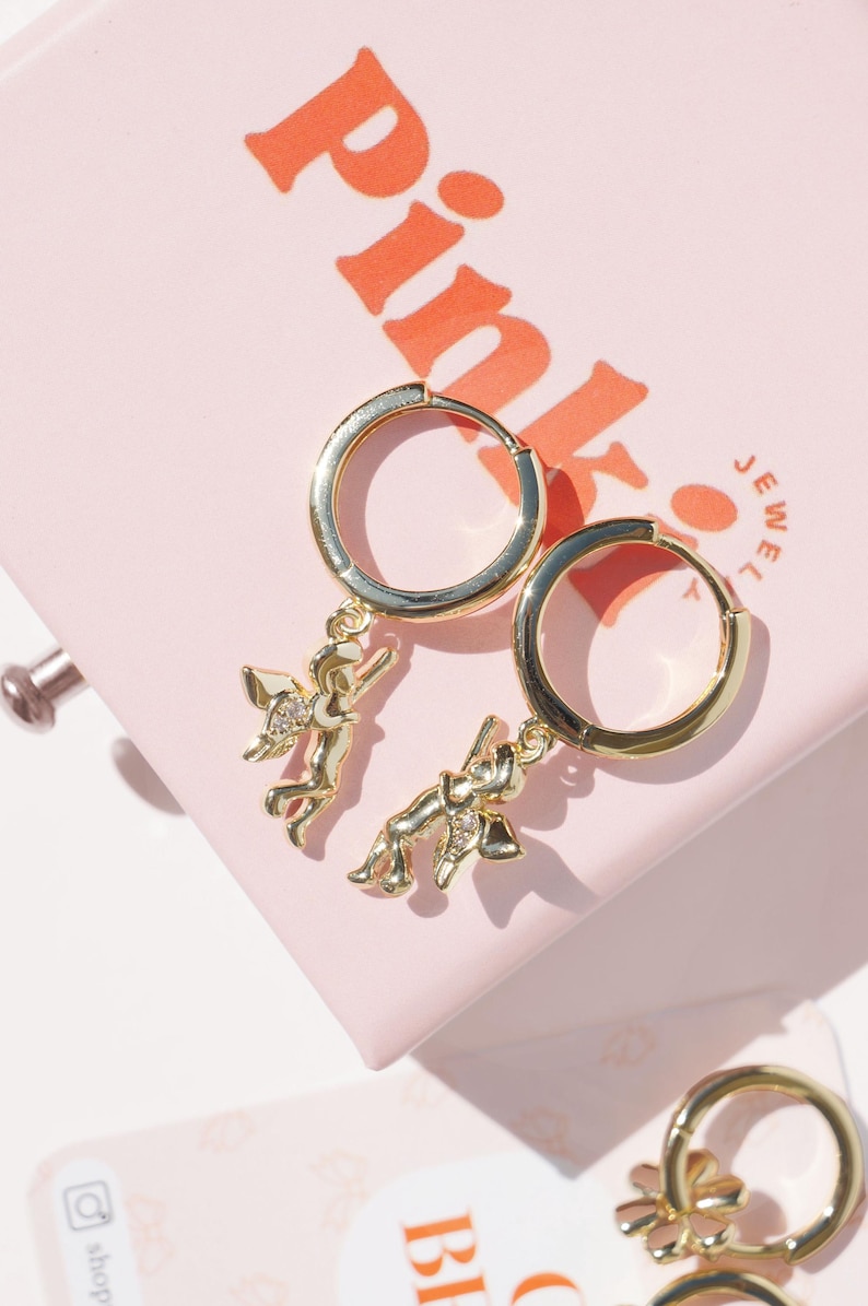 May include: Gold-colored hoop earrings with a charm of a small figure with outstretched arms. The earrings are displayed on a pink surface with the word "Pink" in orange letters.