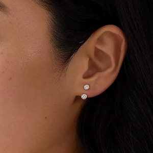 May include: Close-up of two small, round, gold earrings with a single, clear gemstone in each. The earrings are set in a gold metal and are worn on an ear. The ear is set against a blurred background of skin and dark hair.