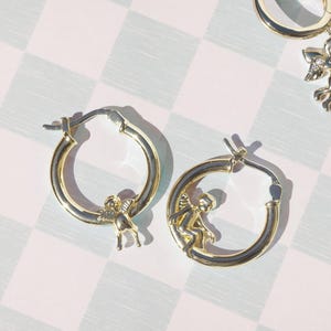 May include: Gold-colored hoop earrings with a small angel charm. The earrings have a hinged closure and a polished finish. The angel charm is detailed with wings and a seated pose. The earrings are displayed on a checkered background.