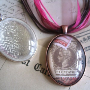 May include: Two oval pendants. One is a silver-toned setting with a textured interior. The other is a copper-toned pendant with a vintage stamp design under glass. The stamp features a profile and the text "3-CENTAVOS". Pink and brown ribbons are attached.
