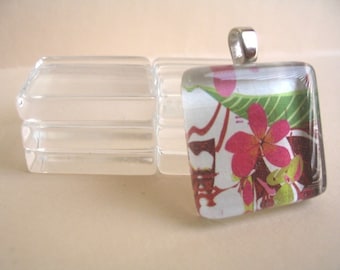 May include: A square glass pendant with a silver bail, featuring a floral design with pink flowers, green leaves, and red accents. Beside it are clear glass rectangular blocks. The pendant is likely for jewelry.