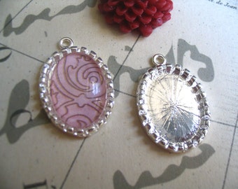 May include: Two silver oval-shaped pendant trays with a decorative border. One tray has a pink background with a white swirl design. The other tray is blank.