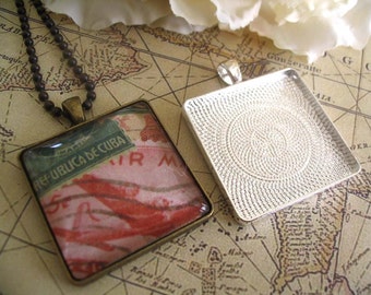 May include: A silver-toned square pendant with a textured surface. The pendant is attached to a black chain. The pendant is lying on a map with the words "Republica de Cuba" visible on a red and white stamp.
