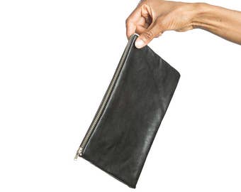 May include: A rectangular black leather clutch bag featuring a smooth texture and a silver-toned metal zipper closure along the top edge. The bag has a simple, slim profile with clean lines and a consistent matte black finish throughout the exterior.