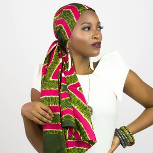May include: A headwrap featuring a vibrant pattern of hot pink and olive green geometric shapes. The fabric is tied securely around the head with a long tail draped over the shoulder. The design includes repeating angular motifs in a bold color scheme.
