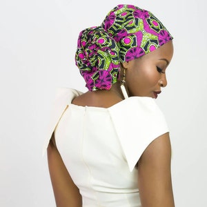 May include: A headwrap featuring a vibrant geometric pattern in shades of magenta, lime green, and black, styled in a voluminous knot at the back. The fabric is tied securely around the head, showcasing a bold, intricate print design.