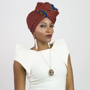 May include: A headwrap featuring a red, blue, and yellow geometric pattern, paired with a white top with structured shoulders. A gold-toned necklace with a circular pendant and long, white, feather-like earrings complete the ensemble.