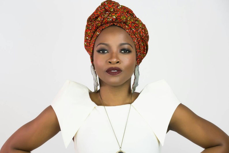 May include: A headwrap featuring a vibrant orange, red, and green patterned fabric, paired with large, elongated, translucent gray earrings and a delicate gold-toned necklace with a circular pendant, worn over a white top with structured shoulders.