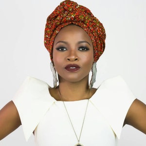 May include: A headwrap featuring a vibrant orange, red, and green geometric pattern, paired with large, elongated, light-colored, textured drop earrings and a simple pendant necklace worn with a solid white top with structured, flared shoulders.