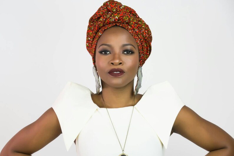 May include: A headwrap featuring a vibrant orange, red, and green geometric pattern, paired with large, elongated, light-colored, textured drop earrings and a simple pendant necklace worn with a solid white top with structured, flared shoulders.