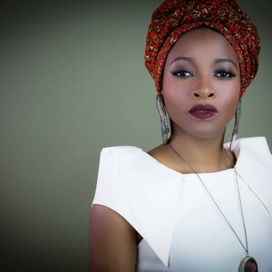 May include: A white sleeveless top with structured shoulders, paired with a red and gold patterned headwrap. The ensemble includes long dangling earrings and a silver-toned pendant necklace featuring a dark circular charm.