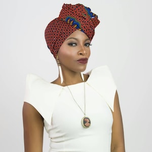 May include: A headwrap featuring a geometric pattern in red, blue, and yellow, paired with a white sleeveless top with structured shoulders. A long gold-tone necklace with a round pendant and long white feather-shaped earrings complete the look.